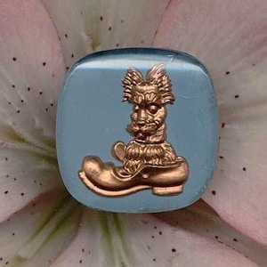 May include: A blue glass cabochon with a gold-toned image of a dog wearing a shoe. The dog is sitting in the shoe and has a playful expression.