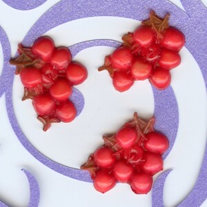 Puede incluir: Tres racimos de uvas de pl&aacute;stico rojo con tallos marrones. Las uvas est&aacute;n dispuestas sobre una superficie blanca con un patr&oacute;n de espiral morado.