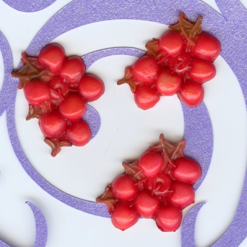 Puede incluir: Tres racimos de uvas de pl&aacute;stico rojo con tallos marrones. Las uvas est&aacute;n dispuestas sobre una superficie blanca con un patr&oacute;n de espiral morado.