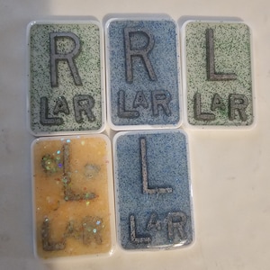 Pode incluir: Cinco peças retangulares de resina com glitter e letras. As letras "R LAR" e "L LAR" são visíveis em prata. Os fundos são verde claro, azul e amarelo. As peças têm bordas brancas.