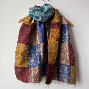 May include: A patchwork silk scarf featuring a range of colours and patterns, including burgundy, gold, blue, and white. The scarf is folded and draped over a wooden hanger.
