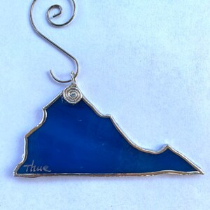May include: A blue glass ornament shaped like the state of Virginia with a silver wire hanger and the word "Thue" written on the bottom.