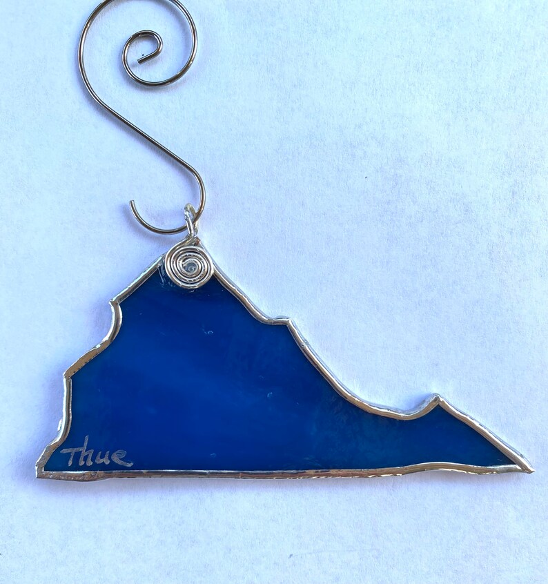 May include: A blue glass ornament shaped like the state of Virginia with a silver wire hanger and the word "Thue" written on the bottom.