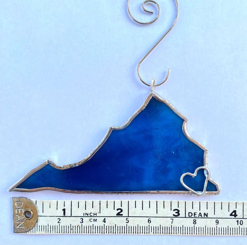 May include: A blue stained glass ornament shaped like the state of Virginia with a small heart in the bottom right corner. The ornament is approximately 3 inches wide.