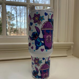Puede incluir: Vaso blanco con un personaje de dibujos animados azul y morado, sosteniendo una bebida morada. El dise&ntilde;o incluye flores, corazones y estrellas. El vaso tiene una tapa transparente y est&aacute; dise&ntilde;ado para bebidas fr&iacute;as.