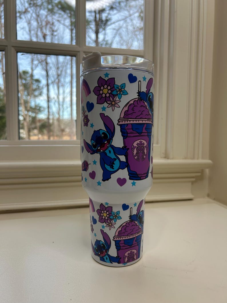 Puede incluir: Vaso blanco con un personaje de dibujos animados azul y morado, sosteniendo una bebida morada. El dise&ntilde;o incluye flores, corazones y estrellas. El vaso tiene una tapa transparente y est&aacute; dise&ntilde;ado para bebidas fr&iacute;as.