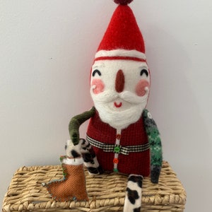 May include: A handmade felt Santa Claus doll with a red hat, white beard, and a green and black patterned outfit. The doll is sitting on a wicker basket and holding a brown boot.
