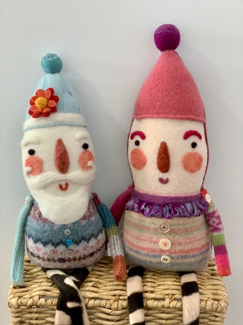 May include: Two handmade felt dolls with striped jumpers and knitted hats. The doll on the left is wearing a blue hat with a flower and a blue and white jumper. The doll on the right is wearing a pink hat and a pink, white and green striped jumper. Both dolls have black and white striped legs and are sitting on a wicker basket.