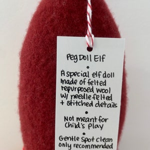 May include: A red felt peg doll elf with a white tag that reads "Peg Doll Elf. A special elf doll made of felted repurposed wool w/ needle felted + stitched details. Not meant for child's play. Gentle Spot clean only recommended made w/ Elf".