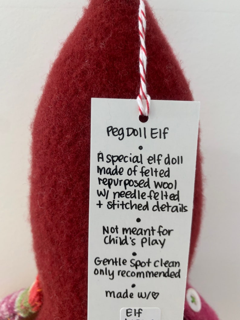 May include: A red felt peg doll elf with a white tag that reads "Peg Doll Elf. A special elf doll made of felted repurposed wool w/ needle felted + stitched details. Not meant for child's play. Gentle Spot clean only recommended made w/ Elf".