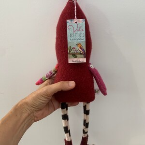 May include: A handmade felt doll with a red body, black and white striped legs, and a purple pom-pom hat. The doll is holding a tag that says "Val's Art Studio" and features a bird on a branch.