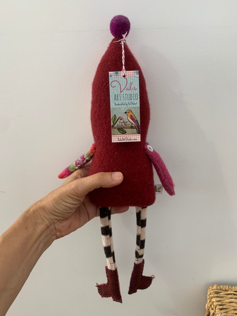 May include: A handmade felt doll with a red body, black and white striped legs, and a purple pom-pom hat. The doll is holding a tag that says "Val's Art Studio" and features a bird on a branch.