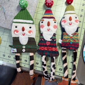 May include: Three handmade felt Christmas decorations shaped like Santa Claus. Each decoration has a different coloured hat and sweater. The decorations have black and white striped legs and are standing on a green and white chequered surface.