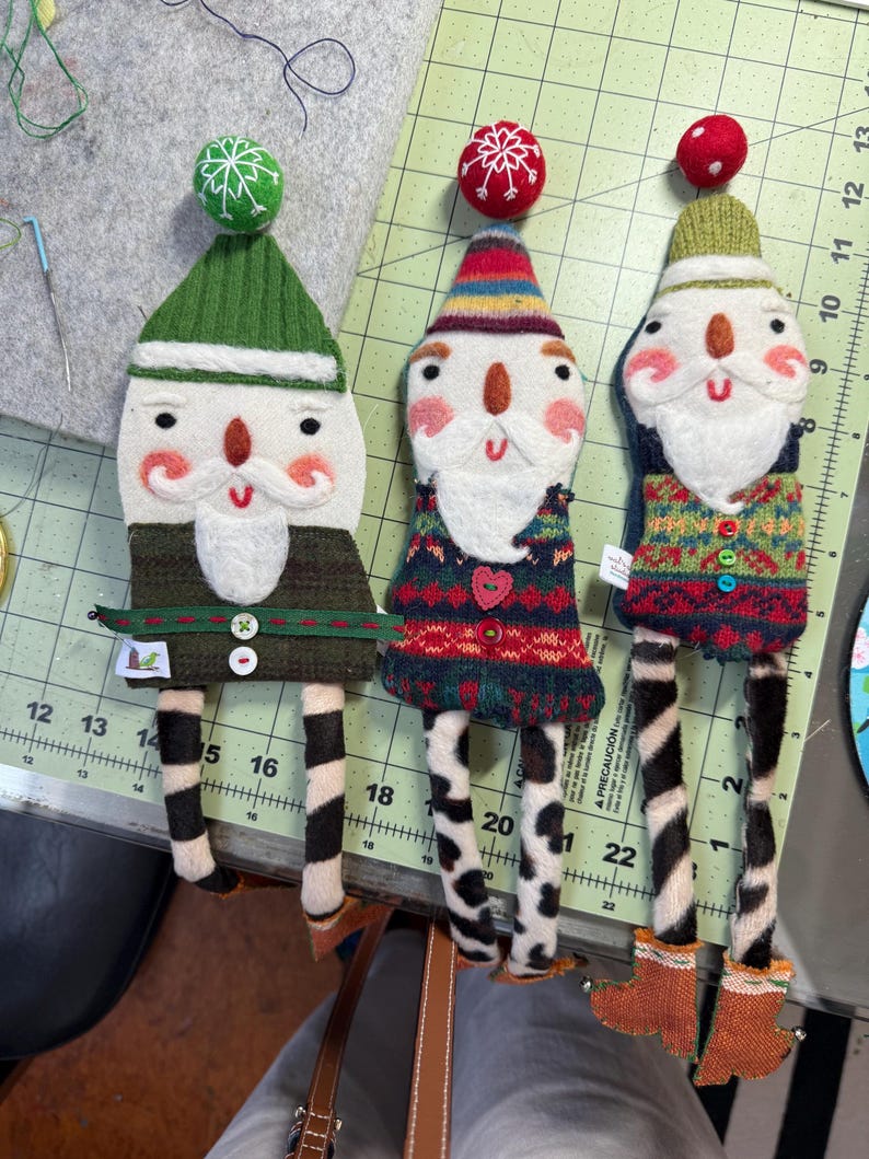 May include: Three handmade felt Christmas decorations shaped like Santa Claus. Each decoration has a different coloured hat and sweater. The decorations have black and white striped legs and are standing on a green and white chequered surface.
