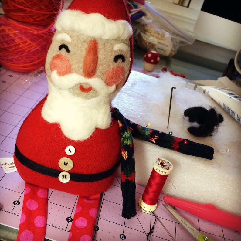 May include: A felt Santa Claus doll with a white beard and red and white suit. The doll is wearing a green and black striped scarf and has red polka dot trousers. The doll is sitting on a pink cutting mat with sewing supplies around it.