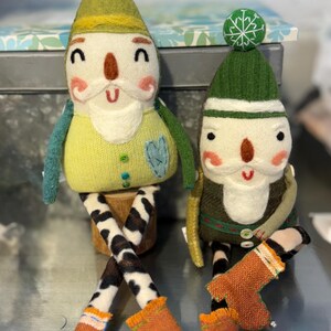 May include: Two handmade felt gnome ornaments with green hats, white beards, and orange boots. The gnomes are sitting on a wooden surface.
