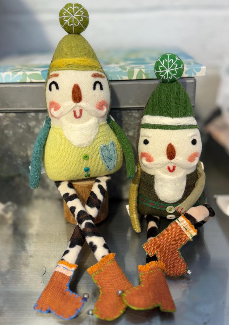 May include: Two handmade felt gnome ornaments with green hats, white beards, and orange boots. The gnomes are sitting on a wooden surface.