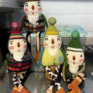 May include: Four handmade felt gnome ornaments with long legs and boots. Each gnome has a different coloured hat and sweater. The gnomes are sitting on a silver metal surface.
