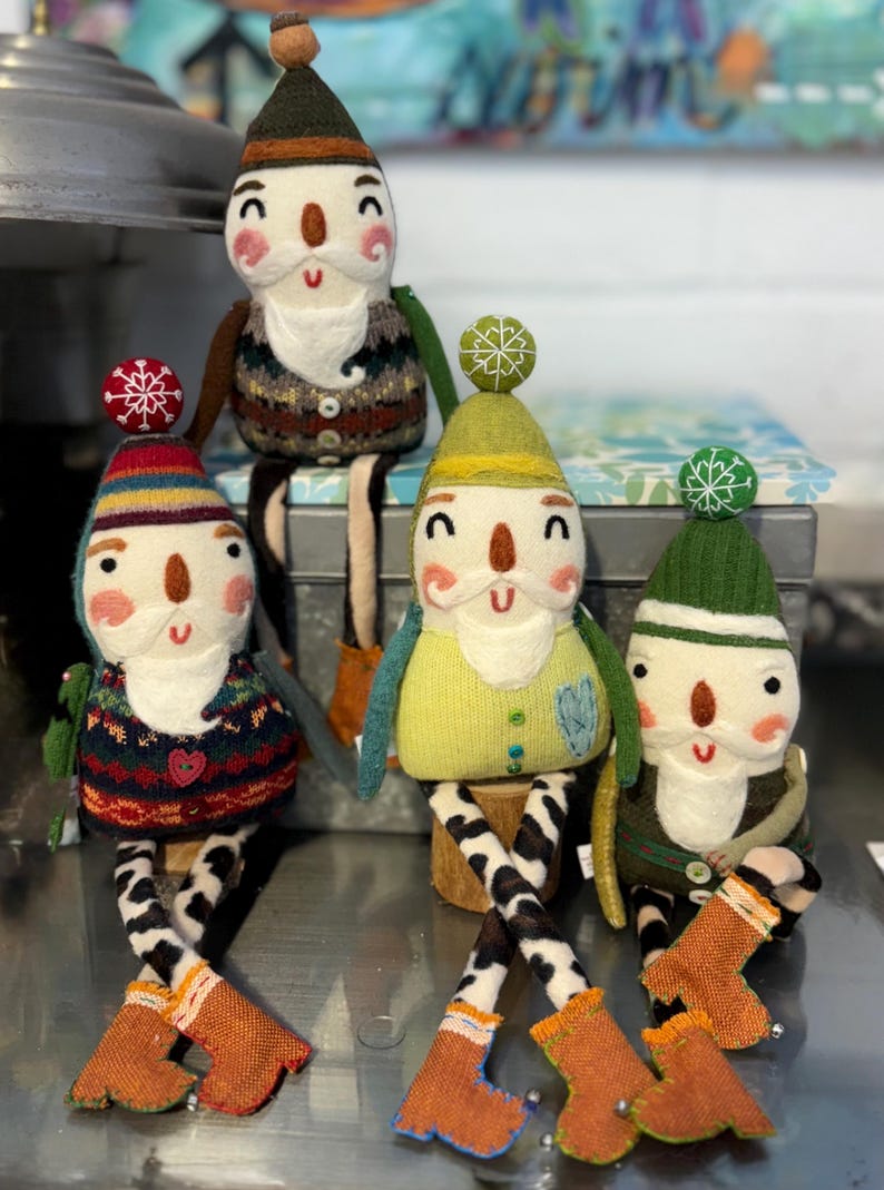 May include: Four handmade felt gnome ornaments with long legs and boots. Each gnome has a different coloured hat and sweater. The gnomes are sitting on a silver metal surface.