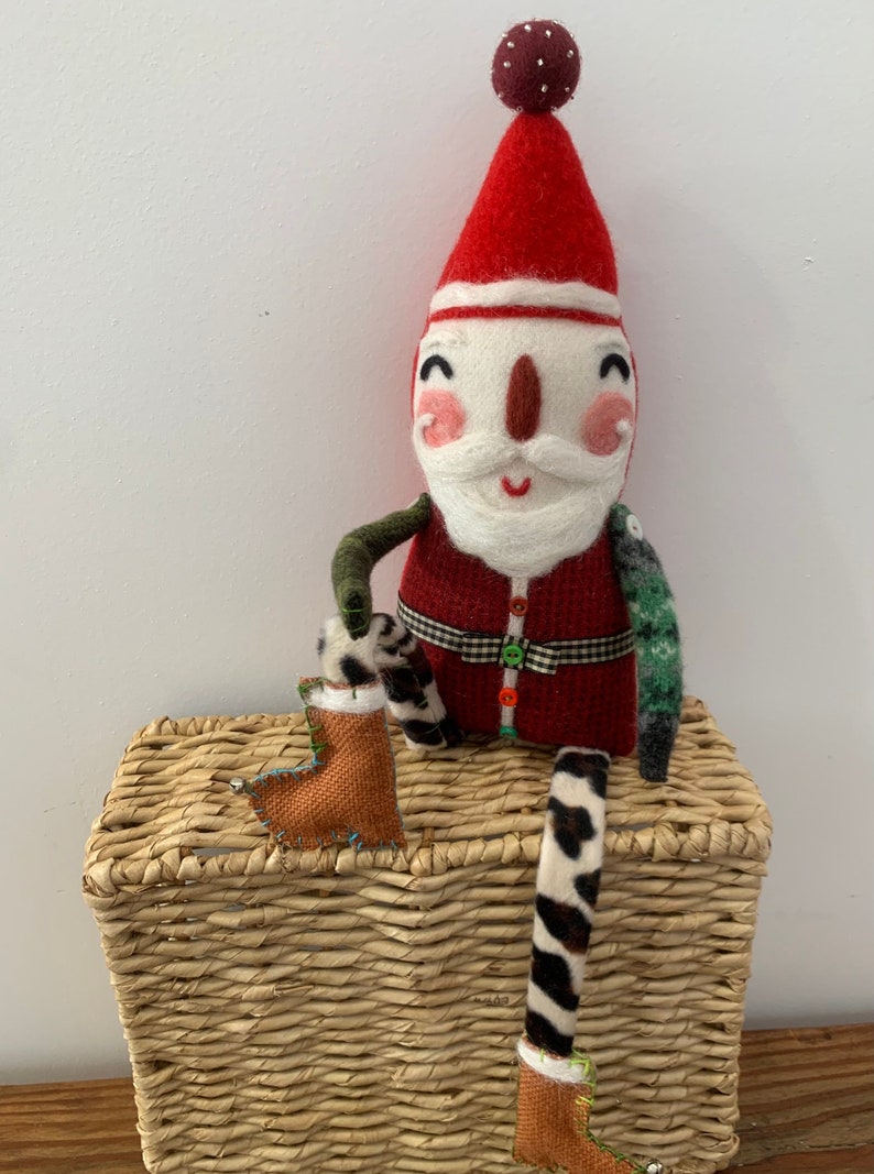 May include: A handmade felt Santa Claus doll with a red hat, white beard, and a green and black patterned outfit. The doll is sitting on a wicker basket and holding a brown boot.