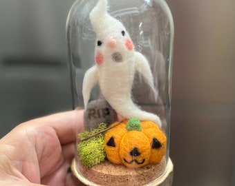 May include: A white felt ghost with pink cheeks sits on a felt pumpkin inside a glass dome. The pumpkin has a carved jack-o'-lantern face. There is a small felt tombstone with the word "RIP" on it.