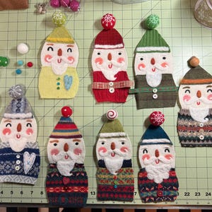 May include: Eight felt ornaments shaped like snowmen with different coloured hats and sweaters. The ornaments are decorated with white felt beards, buttons, and embroidered details.