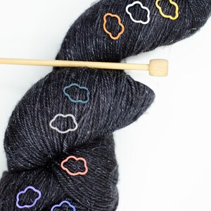 Pode incluir: Uma meada de fio cinza escuro com vários marcadores de ponto coloridos em forma de nuvens. Também é visível uma agulha de tricô de madeira.