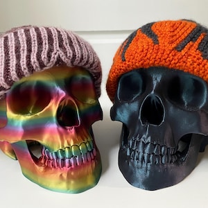 May include: Two colorful skull sculptures wearing knitted hats. One hat is pink and purple striped, the other is orange with gray stripes.