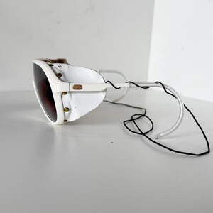 May include: White ski goggles with a leather strap and a brown lens. The goggles are attached to a black cord.