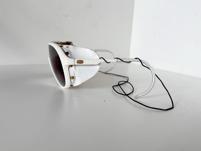 May include: White ski goggles with a leather strap and a brown lens. The goggles are attached to a black cord.