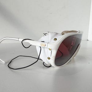 May include: White ski goggles with a leather strap and a dark tinted lens. The goggles have a white frame and a black elastic strap.