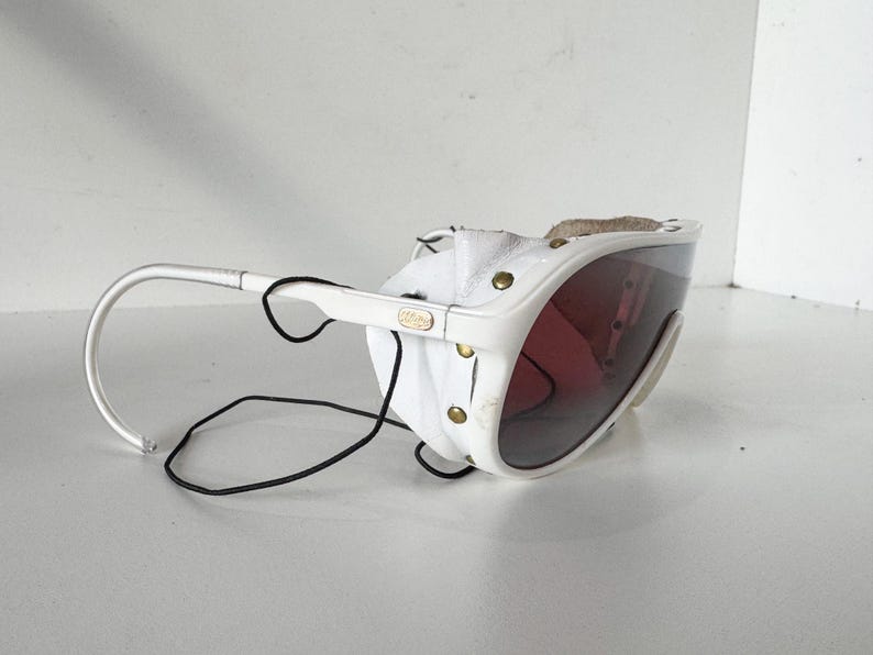 May include: White ski goggles with a leather strap and a dark tinted lens. The goggles have a white frame and a black elastic strap.