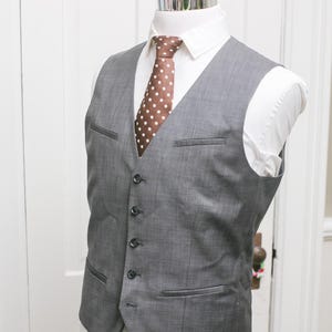May include: A gray vest with a white dress shirt and a brown polka dot tie. The vest has a tailored fit with pockets and buttons. The outfit is displayed on a mannequin.