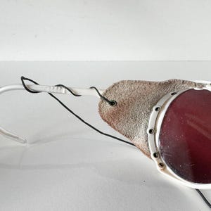May include: White ski goggles with a red lens and a brown leather strap. The goggles have a white frame and a black strap.