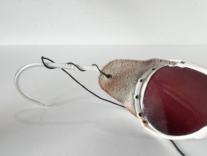 May include: White ski goggles with a red lens and a brown leather strap. The goggles have a white frame and a black strap.