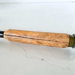 Puede incluir: Primer plano de un mango de madera con un eje de metal oscuro. El mango es de color marr&oacute;n claro con una grieta visible que lo atraviesa. Un anillo y una tapa de metal color lat&oacute;n se encuentran al final del mango.