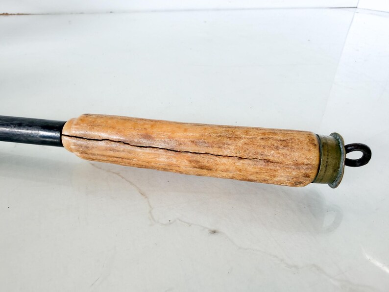 Puede incluir: Primer plano de un mango de madera con un eje de metal oscuro. El mango es de color marr&oacute;n claro con una grieta visible que lo atraviesa. Un anillo y una tapa de metal color lat&oacute;n se encuentran al final del mango.