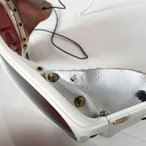 May include: White ski goggles with a leather strap and a white leather nose guard. The goggles have a dark lens and a white frame.