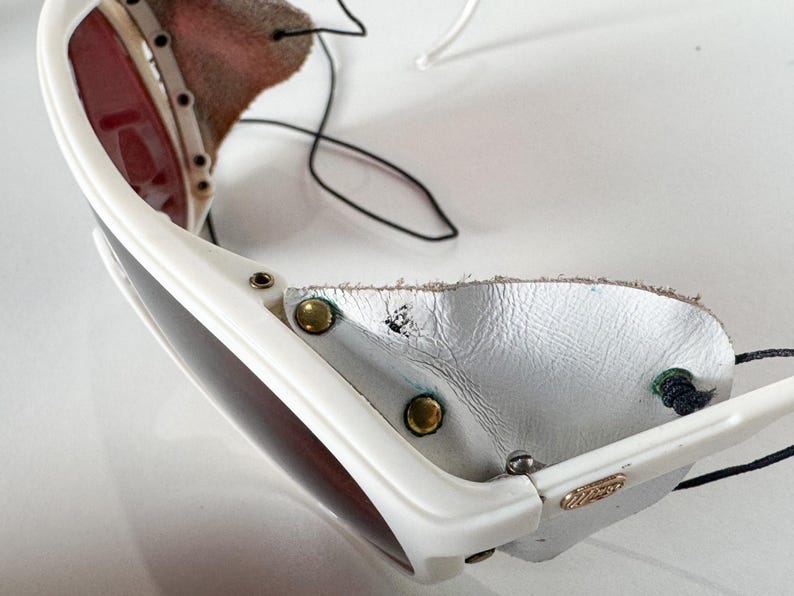 May include: White ski goggles with a leather strap and a white leather nose guard. The goggles have a dark lens and a white frame.
