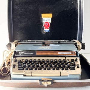 以下が含まれることがあります： オリジナルのケースに入った、ヴィンテージのSCM Smith-Corona Electra 220タイプライター。タイプライターは水色で、黒いキーと木製のアクセントが付いています。ケースは黒と茶色で、「MADE WITH CARE PACKED WITH PRIDE」と書かれたステッカーが付いています。