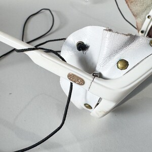 May include: White sunglasses with a black cord and a white leather strap. The sunglasses have a gold logo on the side.