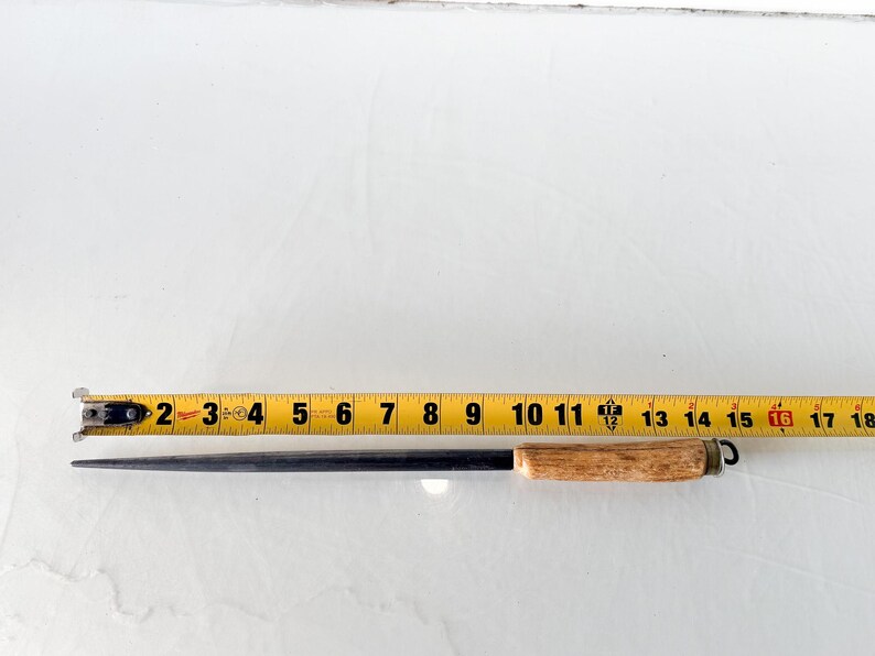 Puede incluir: Un afilador de cuchillos de acero negro con un mango de madera marr&oacute;n claro. El afilador mide aproximadamente 40 cm de largo. Una cinta m&eacute;trica amarilla est&aacute; al lado del afilador.
