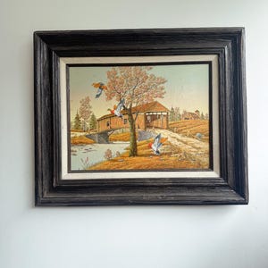 May include: A framed landscape painting showing a covered bridge over a river, with ducks in flight. The artwork has a dark, textured frame and depicts an autumn scene with a tree in full colour.