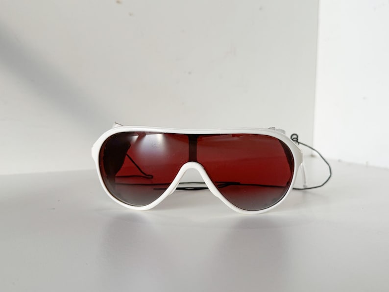 May include: White aviator sunglasses with red tinted lenses. The sunglasses have a white frame and a black strap.