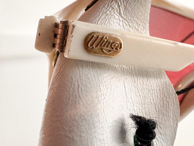 May include: Close-up of a white pair of sunglasses with a gold-colored "Wings" logo on the arm.
