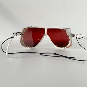 May include: White ski goggles with red lenses and a black strap. The goggles have a white frame and a black strap that goes around the back of the head.