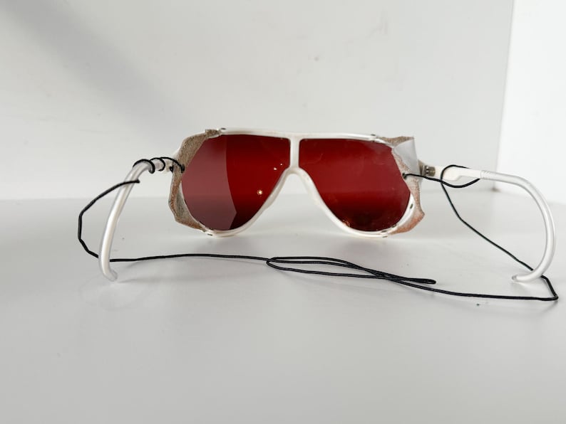 May include: White ski goggles with red lenses and a black strap. The goggles have a white frame and a black strap that goes around the back of the head.