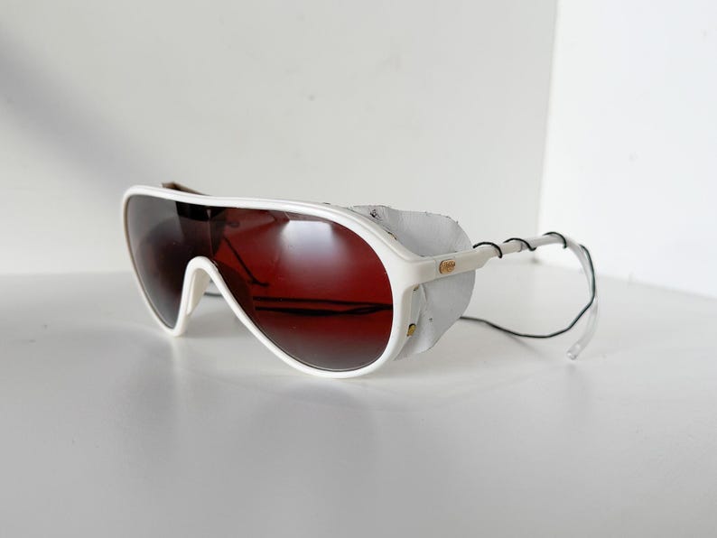 May include: White aviator sunglasses with a red tinted lens and a white leather strap. The sunglasses have a white frame and a black strap.