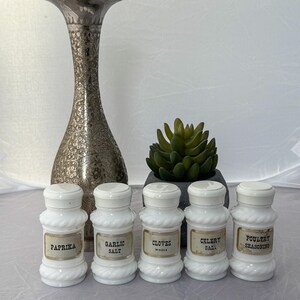 May include: Five white glass spice jars with gold labels. The labels read "Paprika", "Garlic Salt", "Cloves Whole", "Celery Salt", and "Poultry Seasoning".