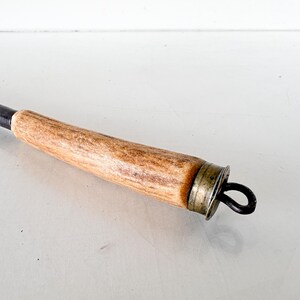 Puede incluir: Primer plano de un atizador de chimenea vintage. El mango es de madera marr&oacute;n claro con un patr&oacute;n texturizado. El extremo met&aacute;lico es oscuro con un lazo para colgar. Un anillo de metal color lat&oacute;n conecta el mango con el atizador.